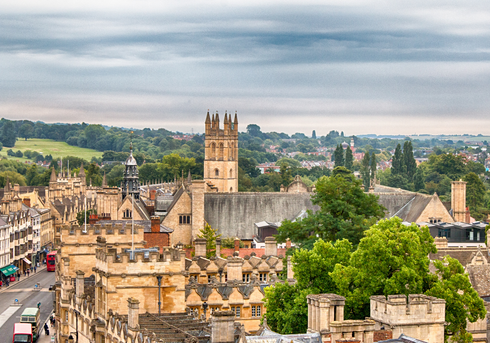 An elevated panoramic view of the historic skyline of Oxford, featuring stone university buildings and Magdalen College Tower beneath an overcast sky.
