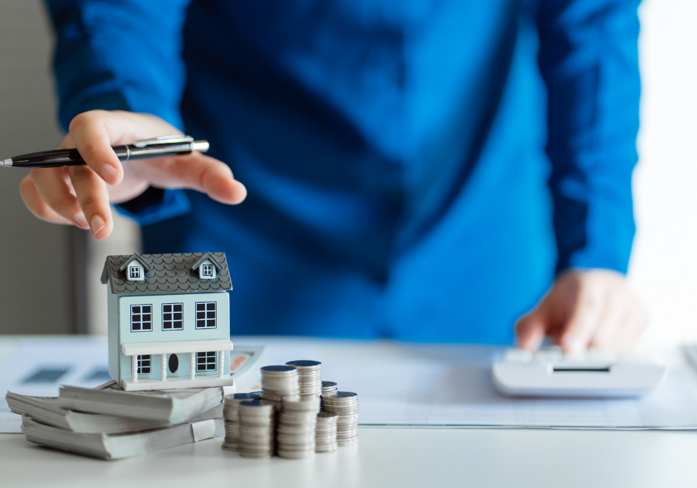 Person using a calculator to compare Sheffield vs Leeds vs Manchester house prices with a miniature home and stacked money.